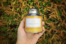 A hand holding a yellow tea can labeled 'Mango Sticky Rice' with a background of green and orange foliage.