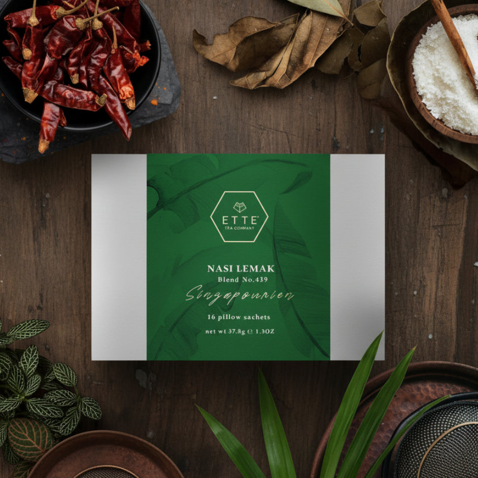 A box of Nasi Lemak tea blend surrounded by dried pandan leaves, dried chilli, and other tea making ingredients, presented on a wooden surface.